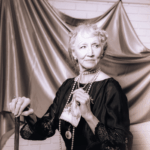 an elegant elderly lady sits in front of a drape holding her cane. Black and white image